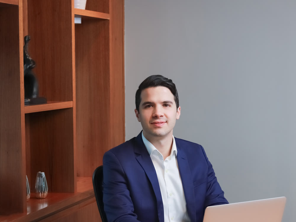 Dr. Danilo Mardegam Razente sentado à mesa do consultório, de terno azul-marinho, diante de um notebook