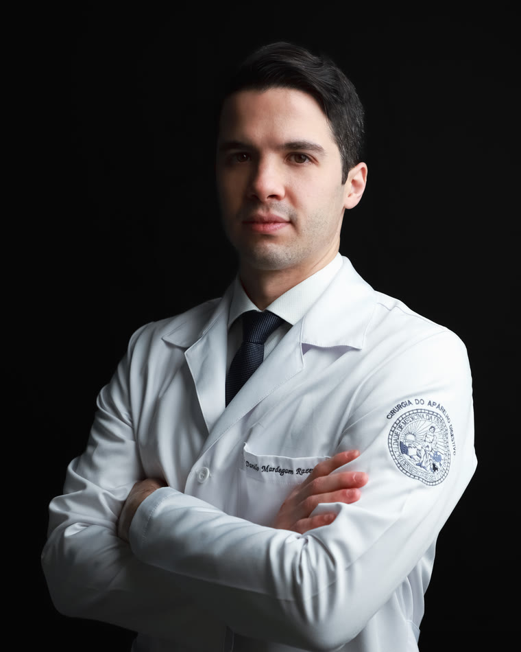 Dr. Danilo Mardegam Razente de jaleco branco com o emblema de Cirurgia do Aparelho Digestivo, braços cruzados